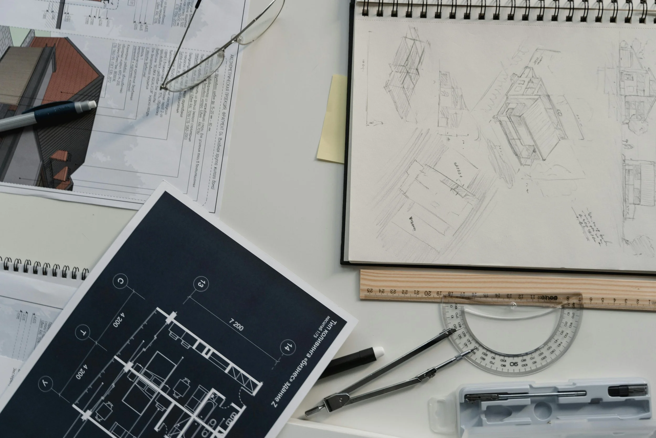 Mesa de trabajo con planos arquitectónicos, una libreta con bocetos, gafas, una regla, un compás, bolígrafos y notas relacionadas con diseño y construcción.