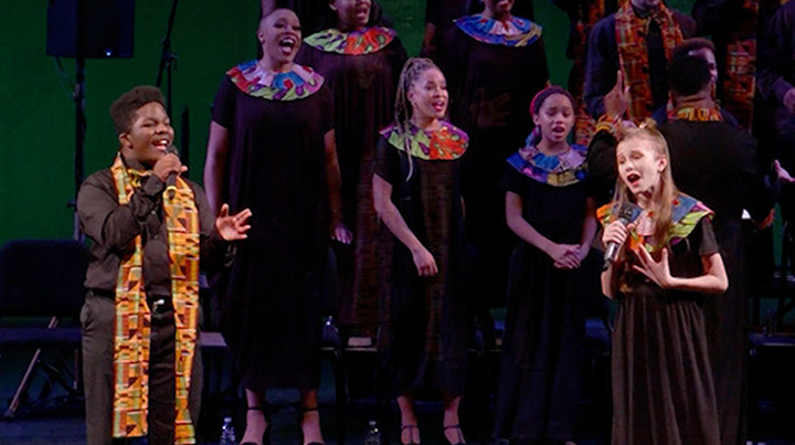 Children singing on stage during a choir or gospel performance, wearing black dresses with colorful necklines and a boy with a patterned sash over his black outfit.