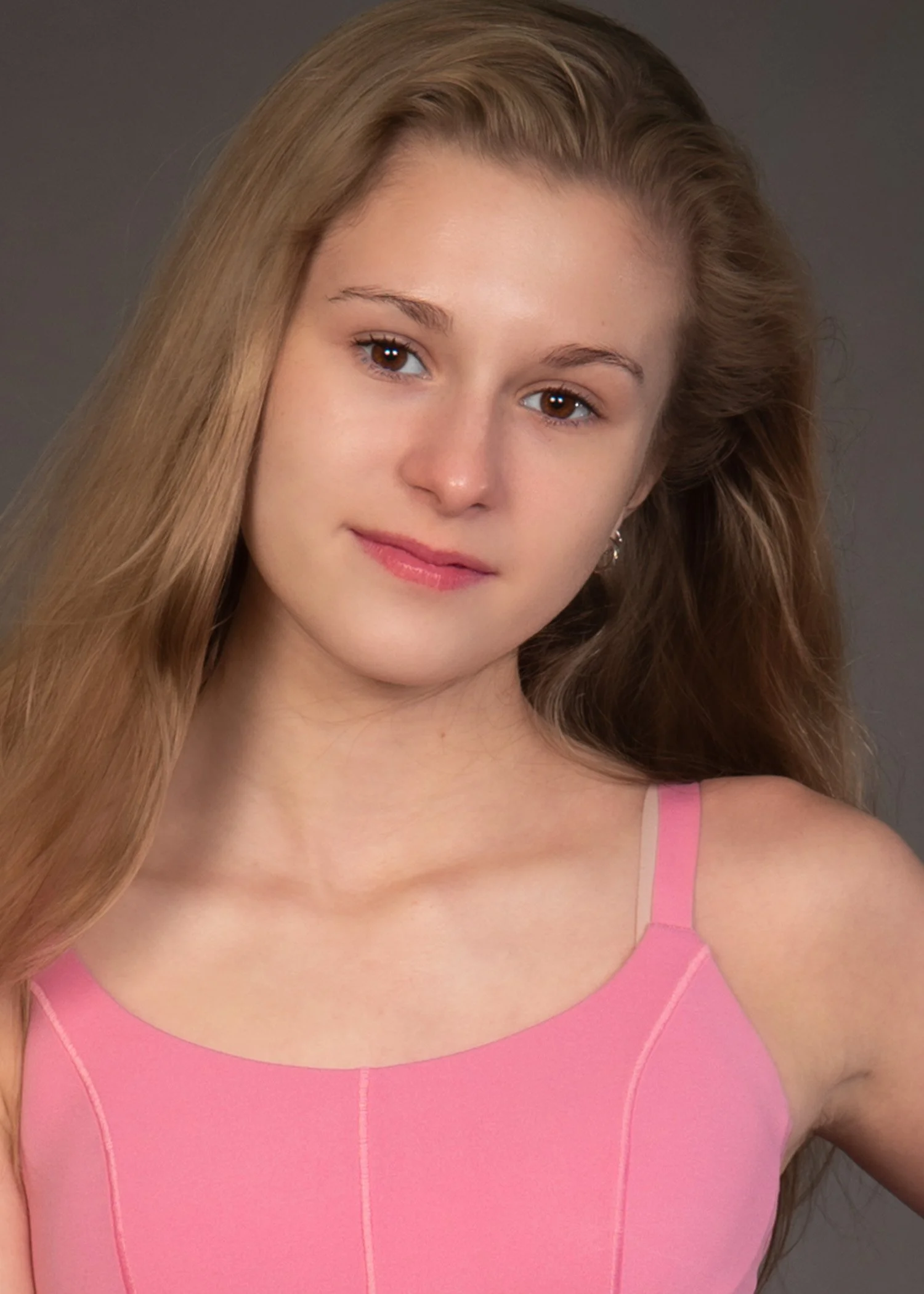 Close-up of a young woman's face with long, wavy dirty blone hair, wearing a pink top with thin straps and smiling gently.