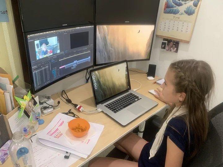 Young girl working on a laptop at a desk with multiple computer monitors, papers, and a calendar in a home office.