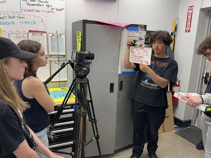 Group of young people in a classroom or studio setting, with one person holding a clapperboard, next to a camera on a tripod, and others watching and preparing for a video or film shoot.