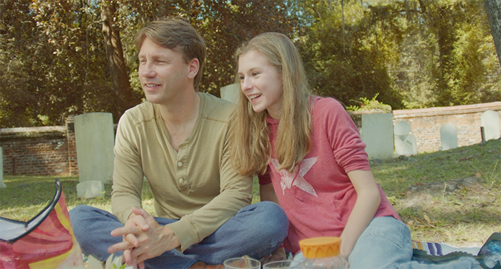 A man and girl sitting on the grass having a picnic, smiling and talking, with trees and a wall in the background.