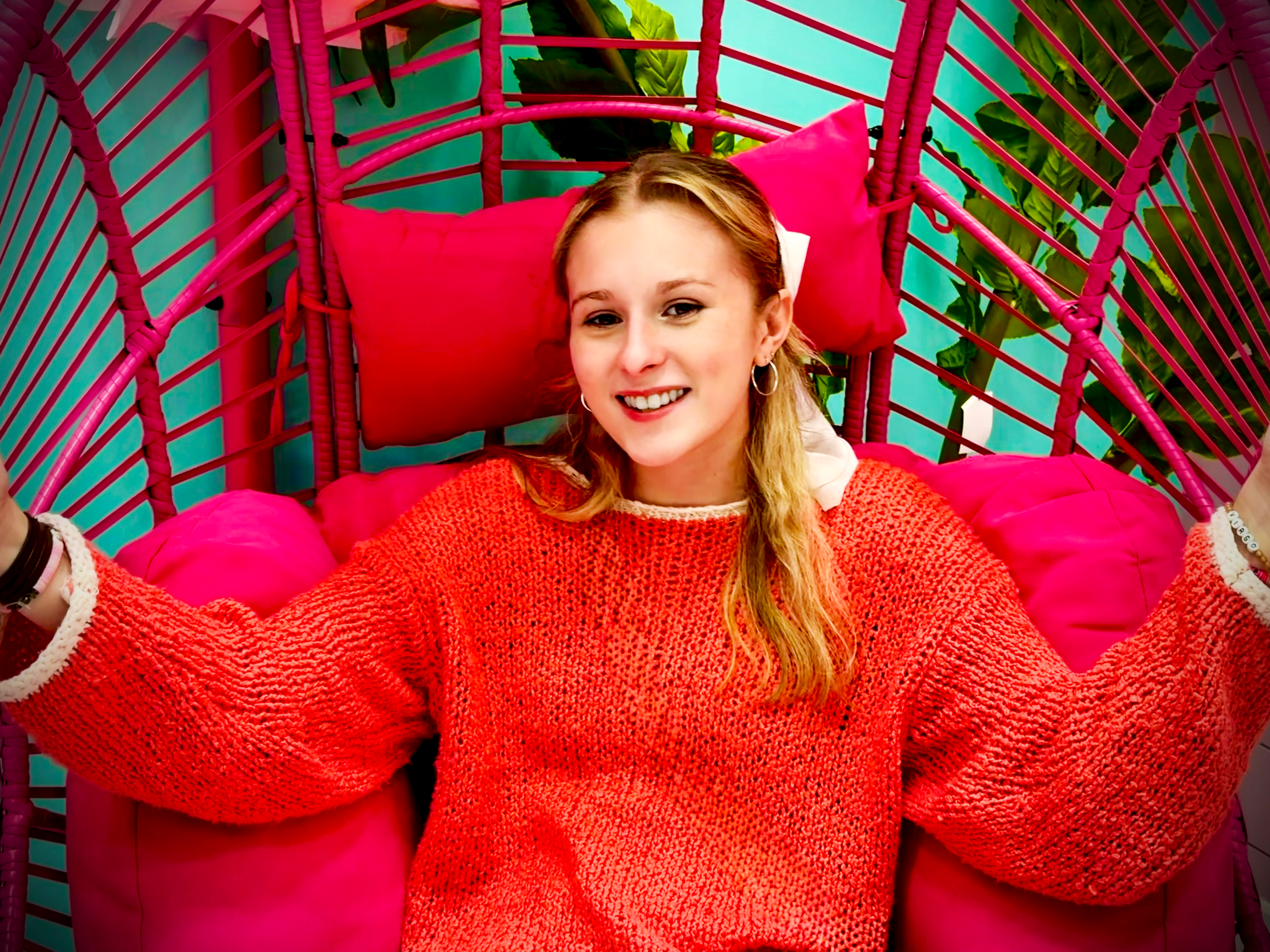 A young woman with dirty blonde hair lying on a pink woven chair with pink cushions, smiling at the camera. The background shows large green leaves and a colorful teal wall.