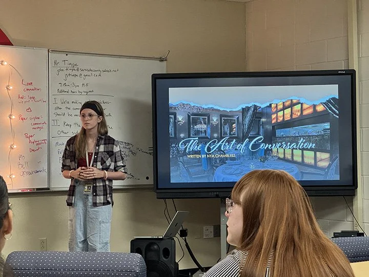 Young girl standing in front of a classroom, giving a presentation titled 'The Art of Conversation' with a slide showing painted windows or murals in an outdoor scene.