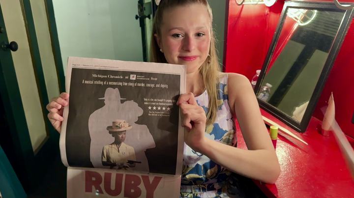 A young girl with blonde hair holding a newspaper with a black-and-white photo and text, sitting at a red counter in a setting resembling a small theater or museum.