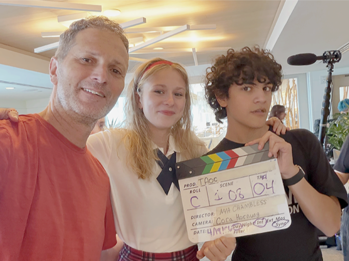 A man and two teenagers holding a movie clapperboard on a film set or studio. The man is wearing a red T-shirt, the girl in a school uniform, and the boy with curly hair in a black T-shirt. Microphone equipment and studio lights are visible in the background.