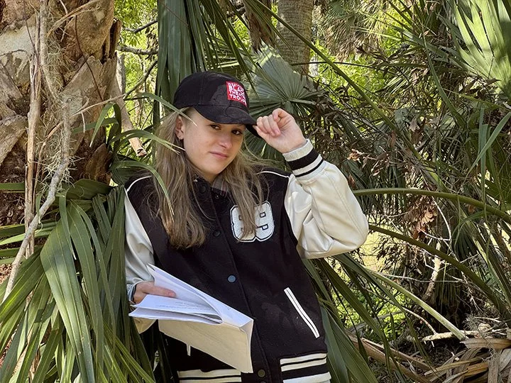 A young woman in a black and white jacket and black cap with a logo, holding papers, standing among jungle-like greenery.