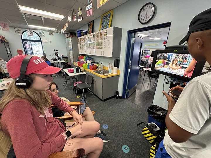 A classroom with students sitting in chairs, one girl wearing a red cap and headphones, and a person filming with a camera. The classroom has educational posters, a clock, and supplies on the counter.
