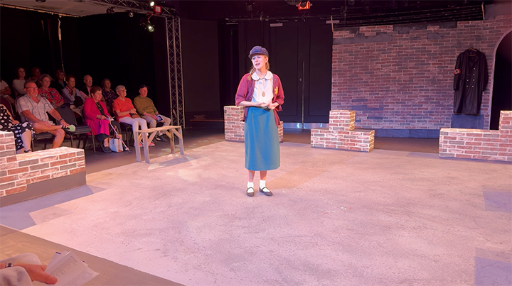 A young actress on stage performing in a theater with an audience seated on the left. The stage has brick walls and minimal props, with the actress dressed in a vintage outfit including a skirt, blouse, and cardigan.