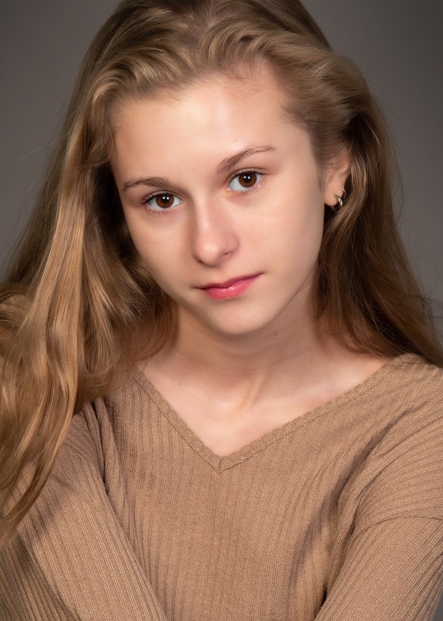 Close-up of a young woman with long, wavy, dirty blonde hair, wearing a tan sweater, looking directly at the camera with a neutral expression.