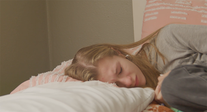A young woman with long blonde hair sleeping peacefully on a bed with white sheets, resting her head on a pillow.
