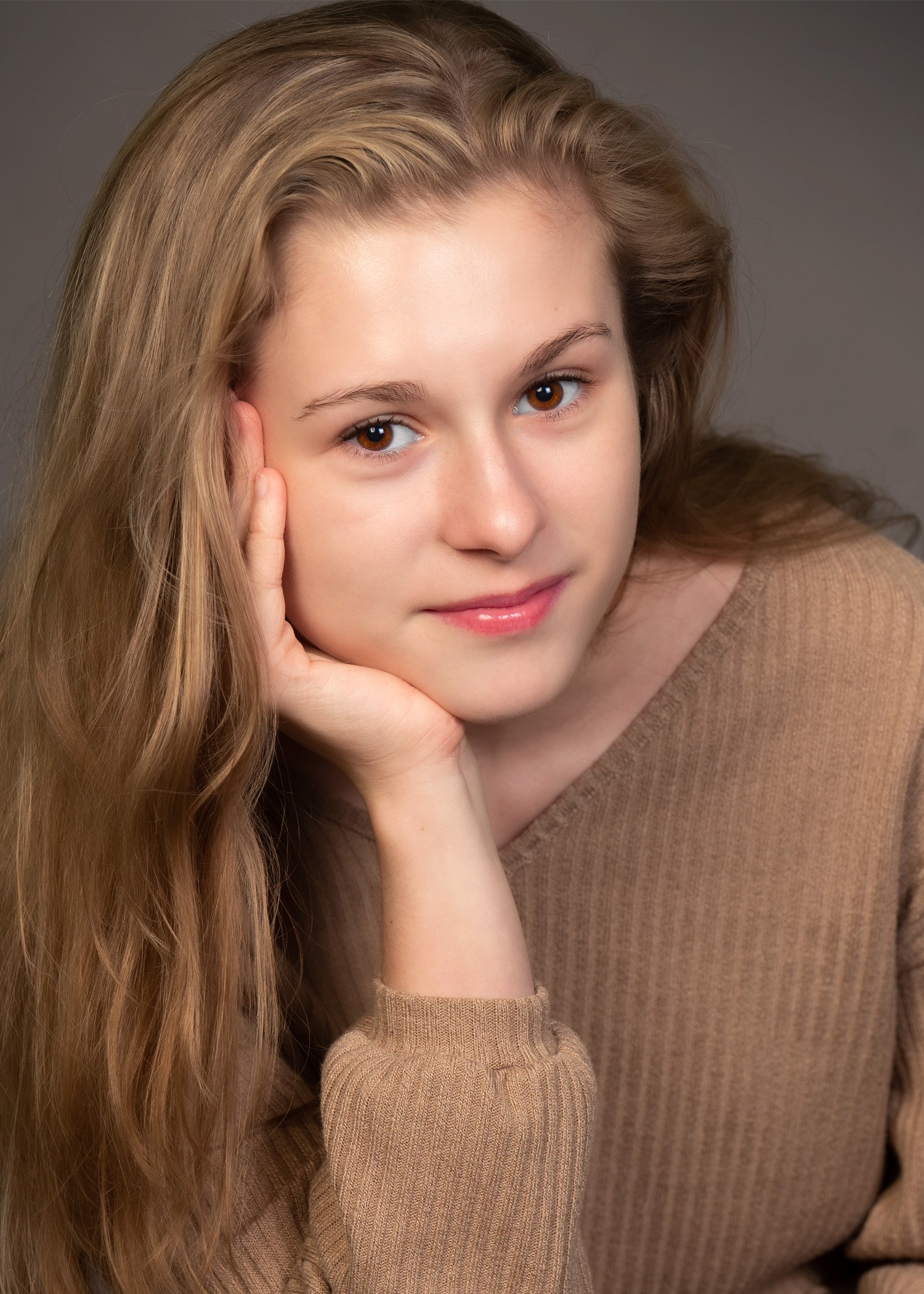 Close-up of a young woman with long, wavy blonde hair, resting her face on her hand, wearing a brown sweater, with a neutral background.