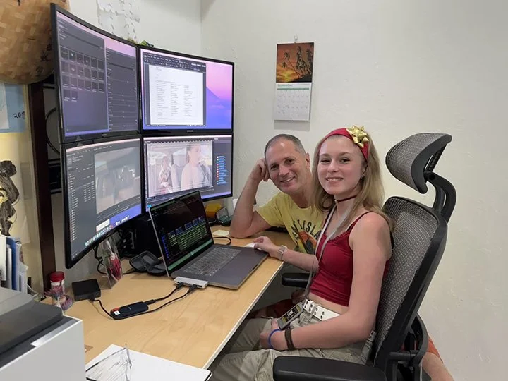 A man and a girl sitting at a desk with multiple computer monitors, working on video editing or digital media projects.