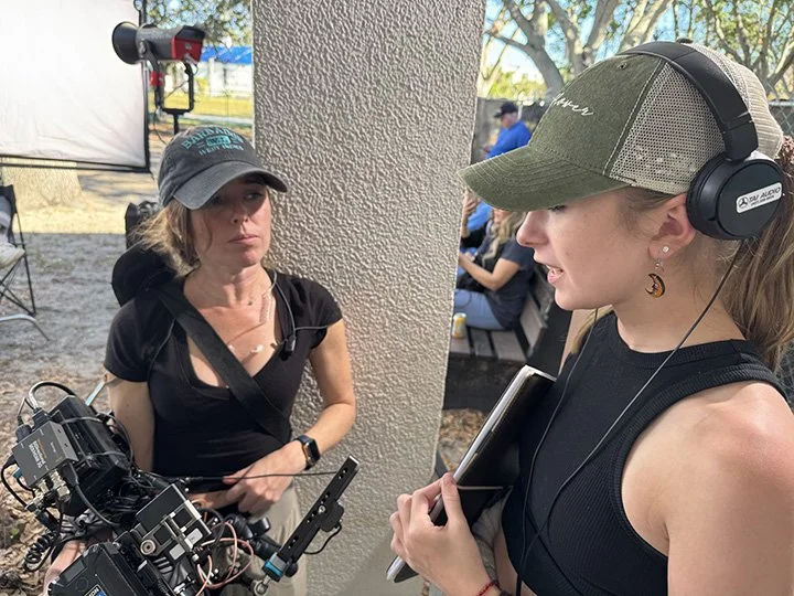 Two women talking outdoors; one woman wearing headphones and holding a tablet, the other woman wearing a cap and standing near camera equipment.