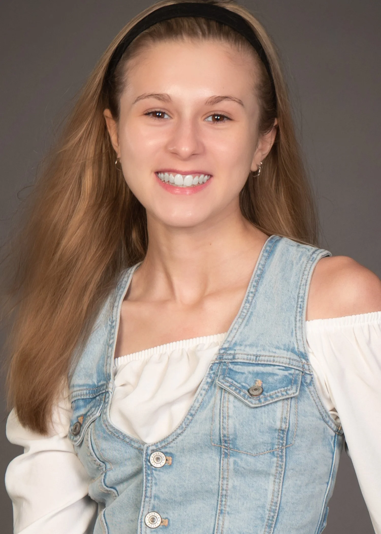 A young woman with long, dirty blonde hair wearing a black headband, a white off-shoulder blouse, and a light blue denim vest smiling at the camera.