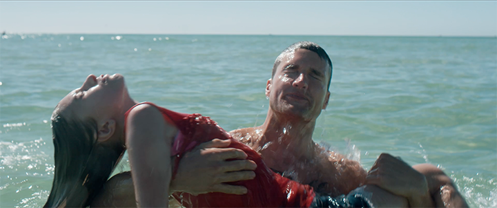 A man is holding a woman in the ocean, with the water and sky in the background.