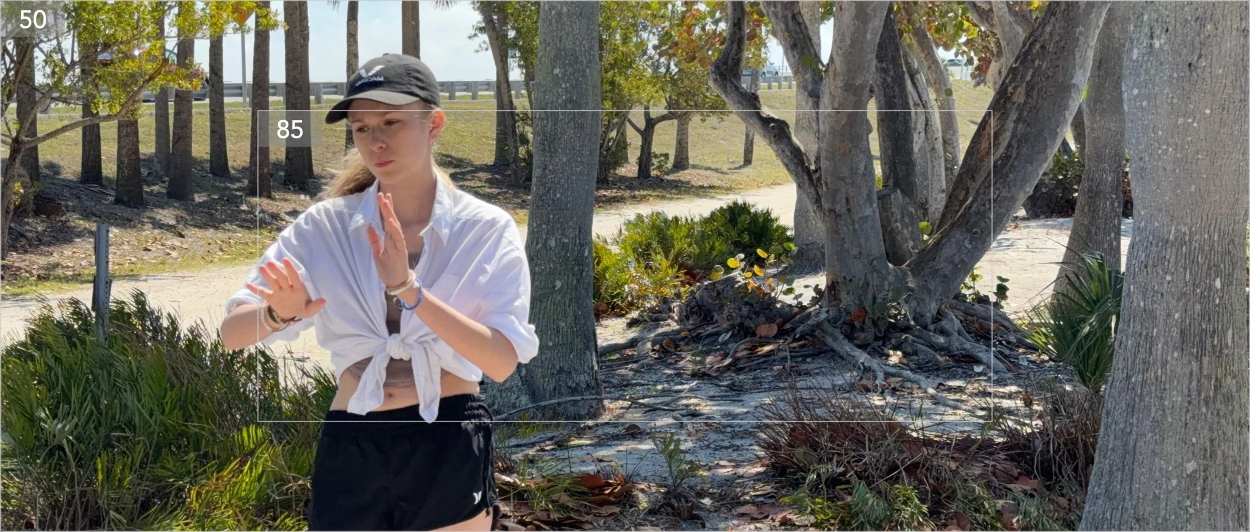 A young woman wearing a black cap, white tied shirt, and black shorts standing outdoors near trees and bushes, with a sandy ground and a distant view of a road and a vehicle.