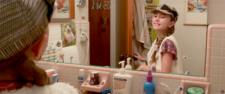 A girl with braided hair wearing a cap and a pink floral shirt, smiling at her reflection in a bathroom mirror, surrounded by toiletries and decorations.