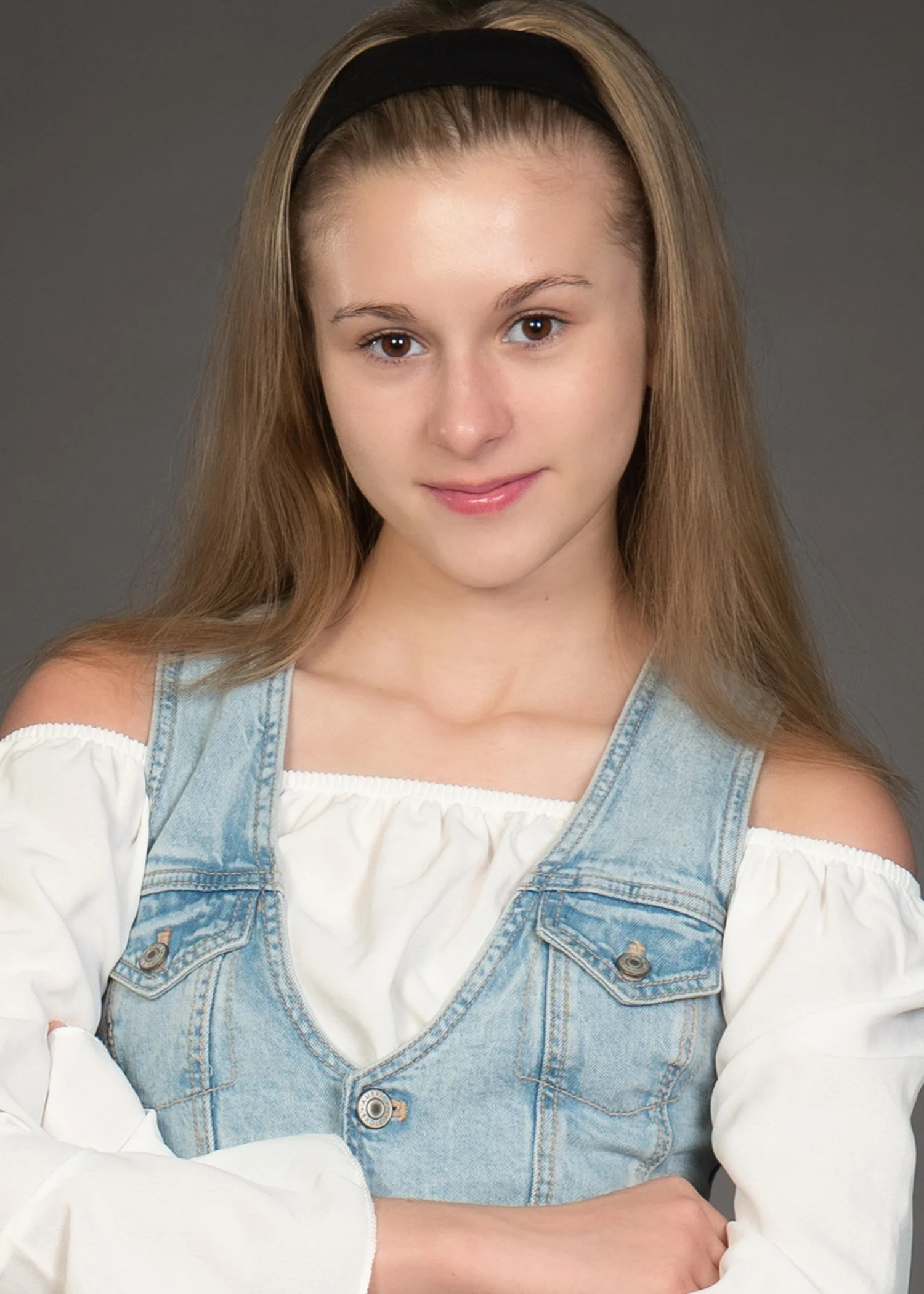 A young woman with long dirty blonde hair, wearing a black headband, denim vest over a white off-shoulder top, smiling slightly, with arms crossed, standing against a gray background.
