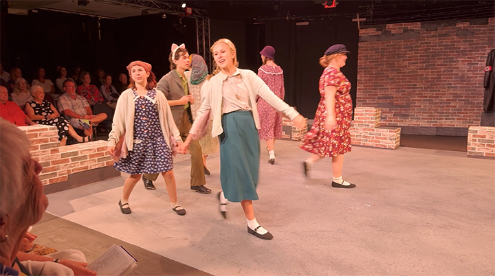Theater performance of women in vintage clothing and hats performing on stage, with audience watching.
