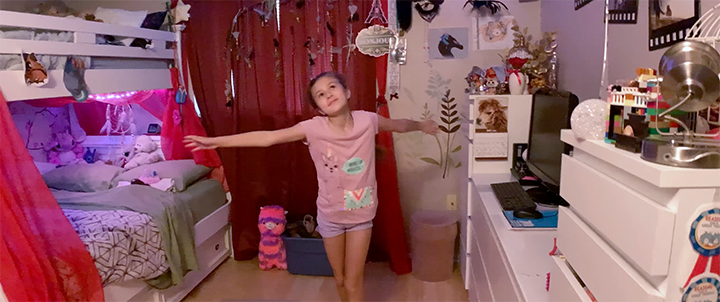 A young girl in a pink shirt and purple shorts stands with arms outstretched in her pink and purple decorated bedroom, with bunk beds, posters, and various toys and decorations.