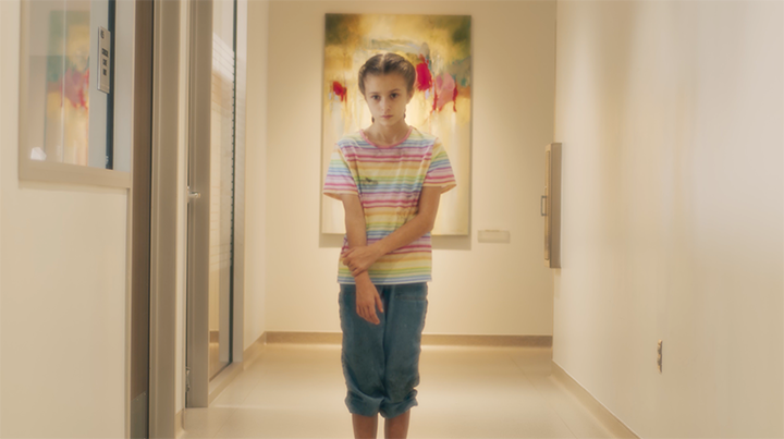 A young girl standing in an art gallery, wearing a colorful striped T-shirt and blue jeans, with artwork hanging on the wall behind her.