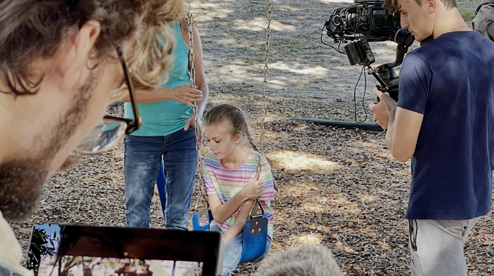 Filming a girl sitting on a swing in a wooded park with a camera crew and director nearby.