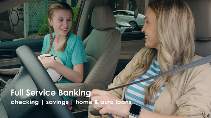A woman and a girl in a car, smiling at each other. The girl is holding a soccer ball and the woman is wearing a seatbelt with a tablet on her wrist. Text overlay says 'Full Service Banking checking | savings | home & auto loans.'