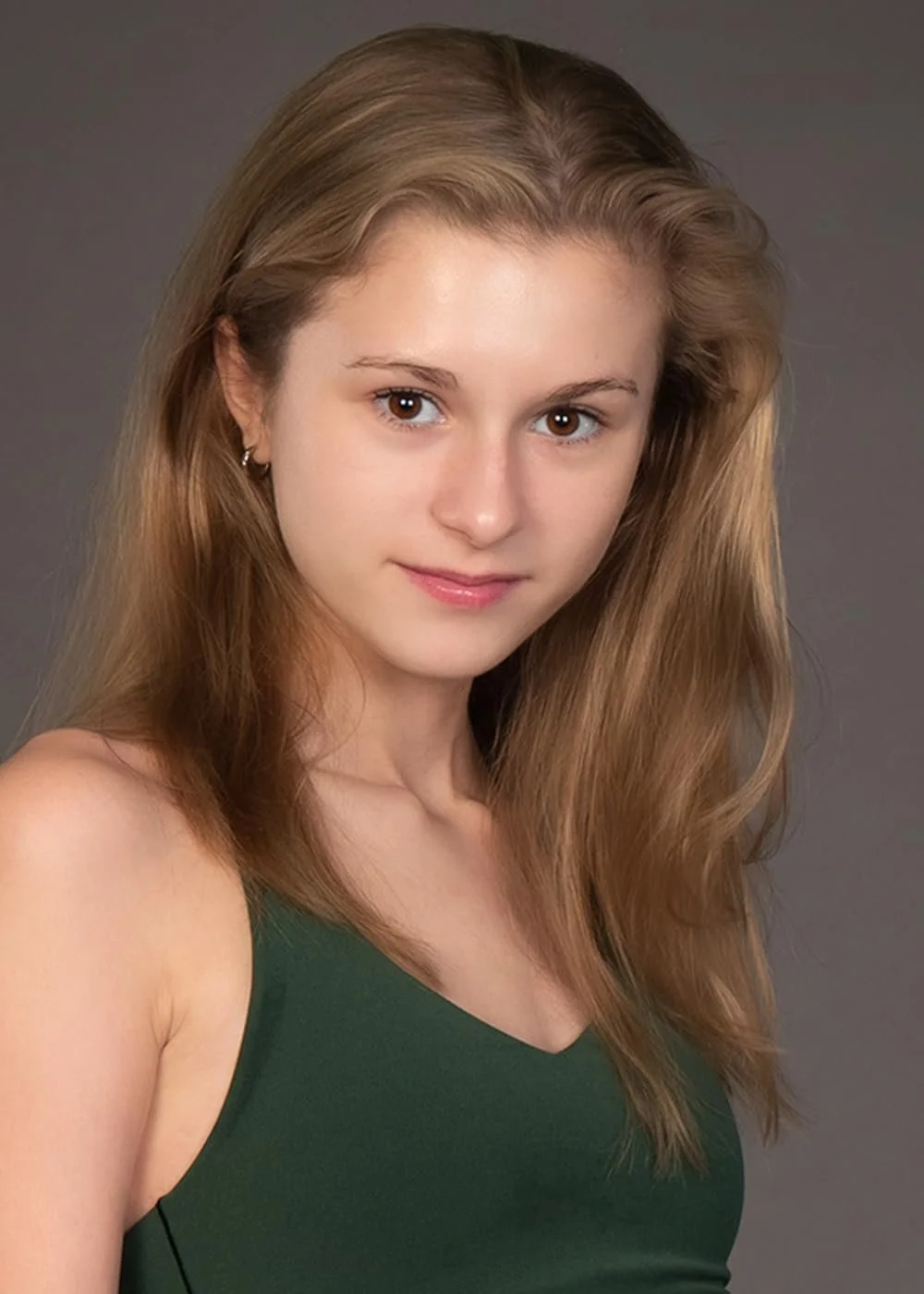 A young woman with long, wavy dirty blonde hair, brown eyes, and fair skin, wearing a sleeveless green top, looking at the camera with a slight smile against a gray background.