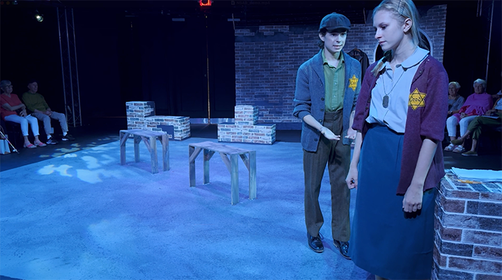Two young actors rehearsing on a stage with brick wall backdrops, chairs, and benches, while a seated audience watches in the background.