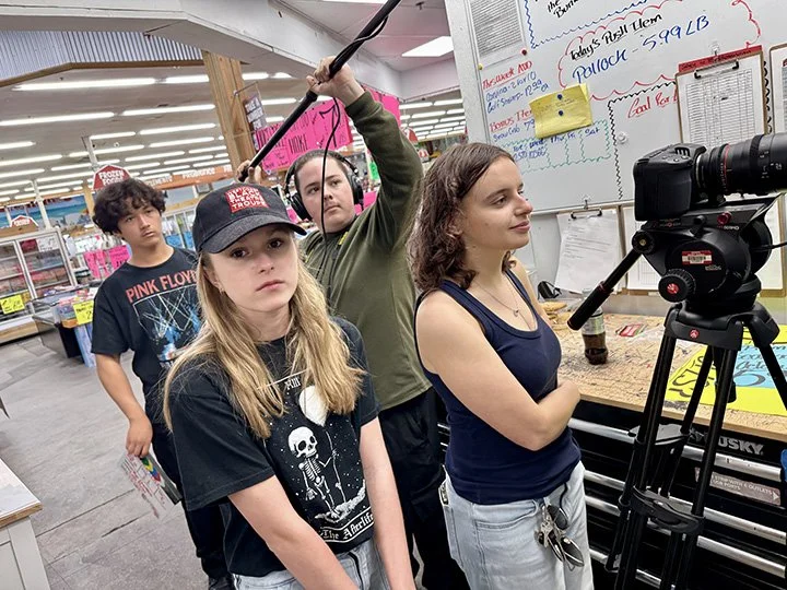 Four teenagers in a store, with one girl operating a camera on a tripod, two others holding a boom microphone, and one standing behind them. They are standing in front of a whiteboard and shopping aisles.