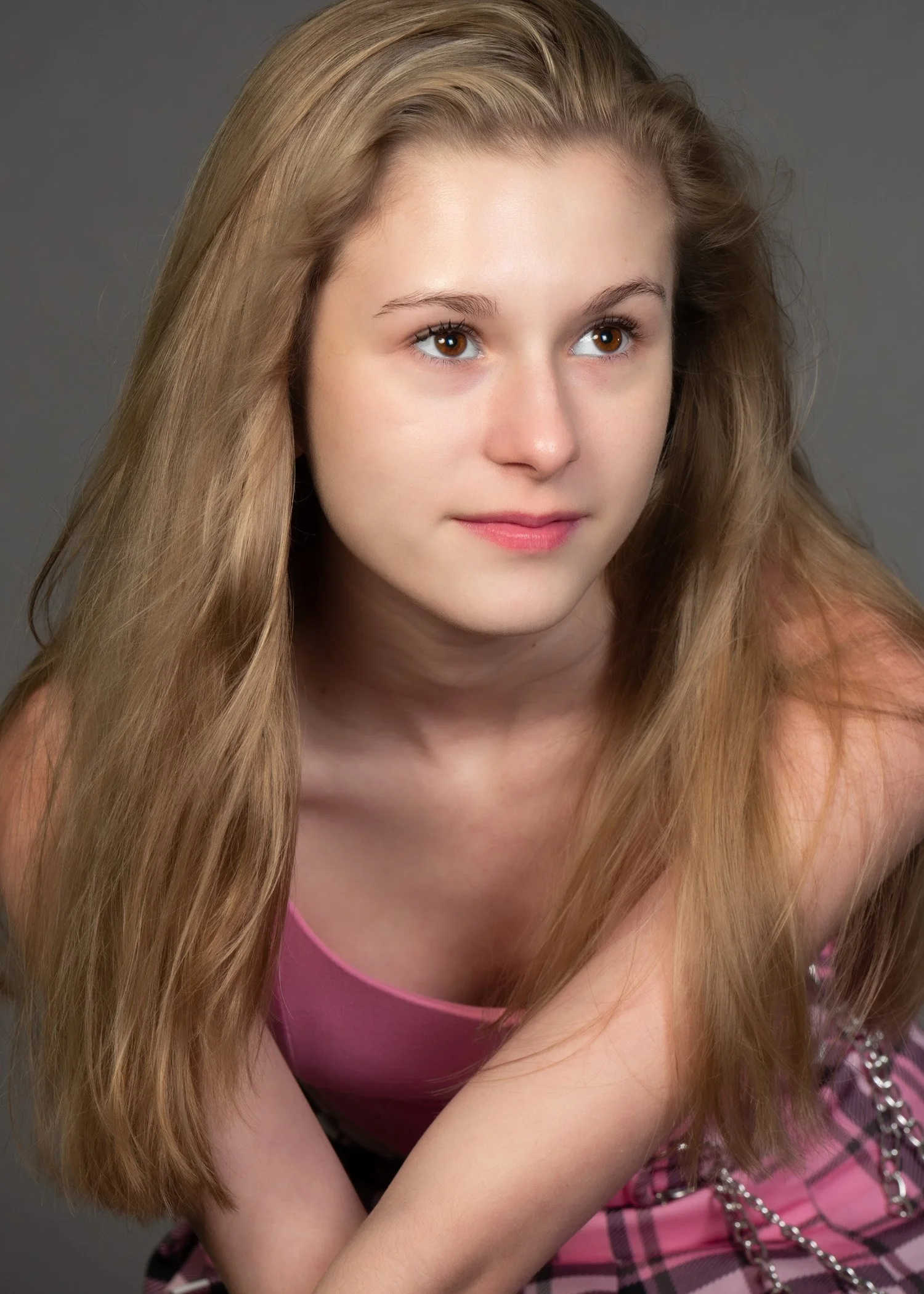 Close-up of a young woman with long, blonde hair, wearing a pink top and a pink plaid skirt with silver chain details.