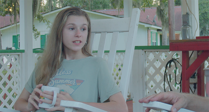 A young girl with long blonde hair sitting outside on a porch, holding a mug, and talking to someone off-camera.