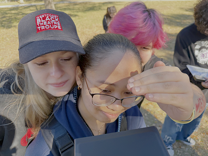 A group of four young people outdoors, closely gathered around a tablet or phone, with sunlight casting shadows on their faces.