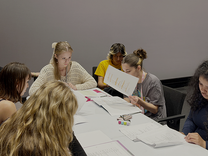 Group of six young women sitting around a table, studying and reviewing documents in a meeting or classroom setting.