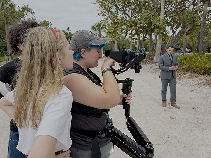 A woman operating a camera on a stabilizer with two young women observing, while a man in a gray suit stands in the background on a sandy outdoor area with trees.