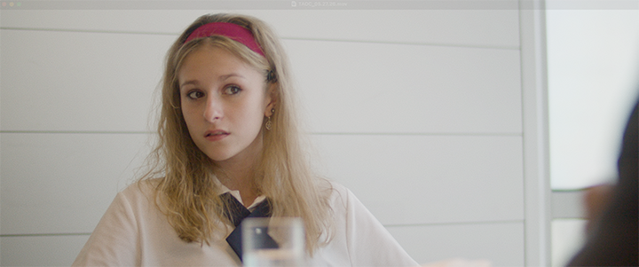 Young woman with blonde hair, pink headband, wearing a beige sweater, sitting at a table with a glass of water in front of her, in a room with white walls.