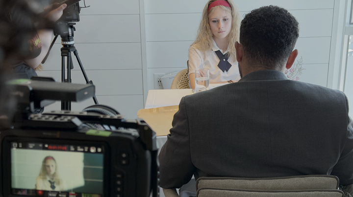 A young girl sits at a table during an interview, facing an adult man. The girl has blond hair with a red headband, wearing a school uniform. A camera records the scene, with the girl visible on the camera screen. The setting is a white-walled room.