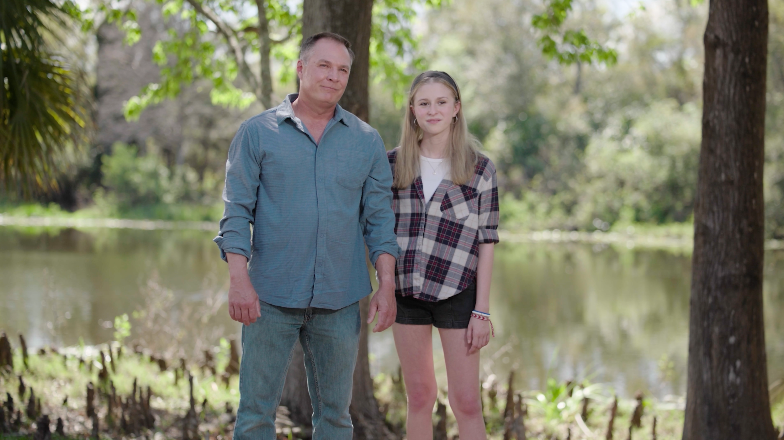 A man and a teenage girl standing outdoors near a body of water in a forested area on a sunny day.