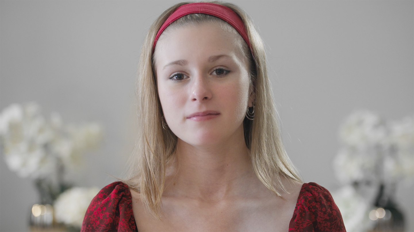 A young woman with blond hair, wearing a red headband and a red patterned top, standing in front of a gray background with blurred white flowers on either side.