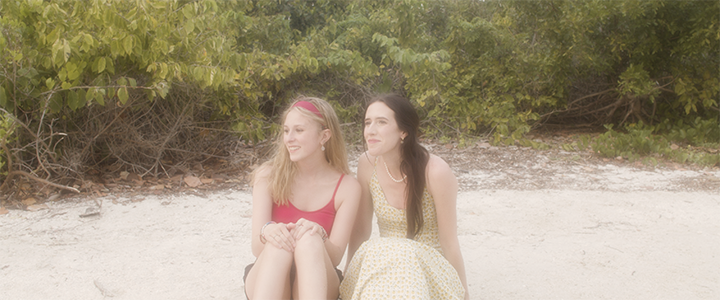 a mother and daughter sitting on a sandy beach near green bushes, enjoying the outdoors.