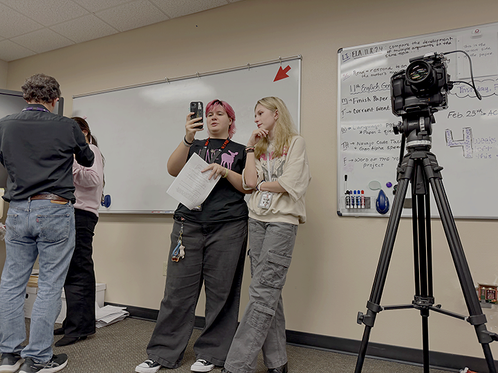 Two young women standing together in a classroom, one with pink hair holding a phone and paper, and the other with blonde hair listening; a camera on a tripod recording them; a whiteboard and teacher in the background.