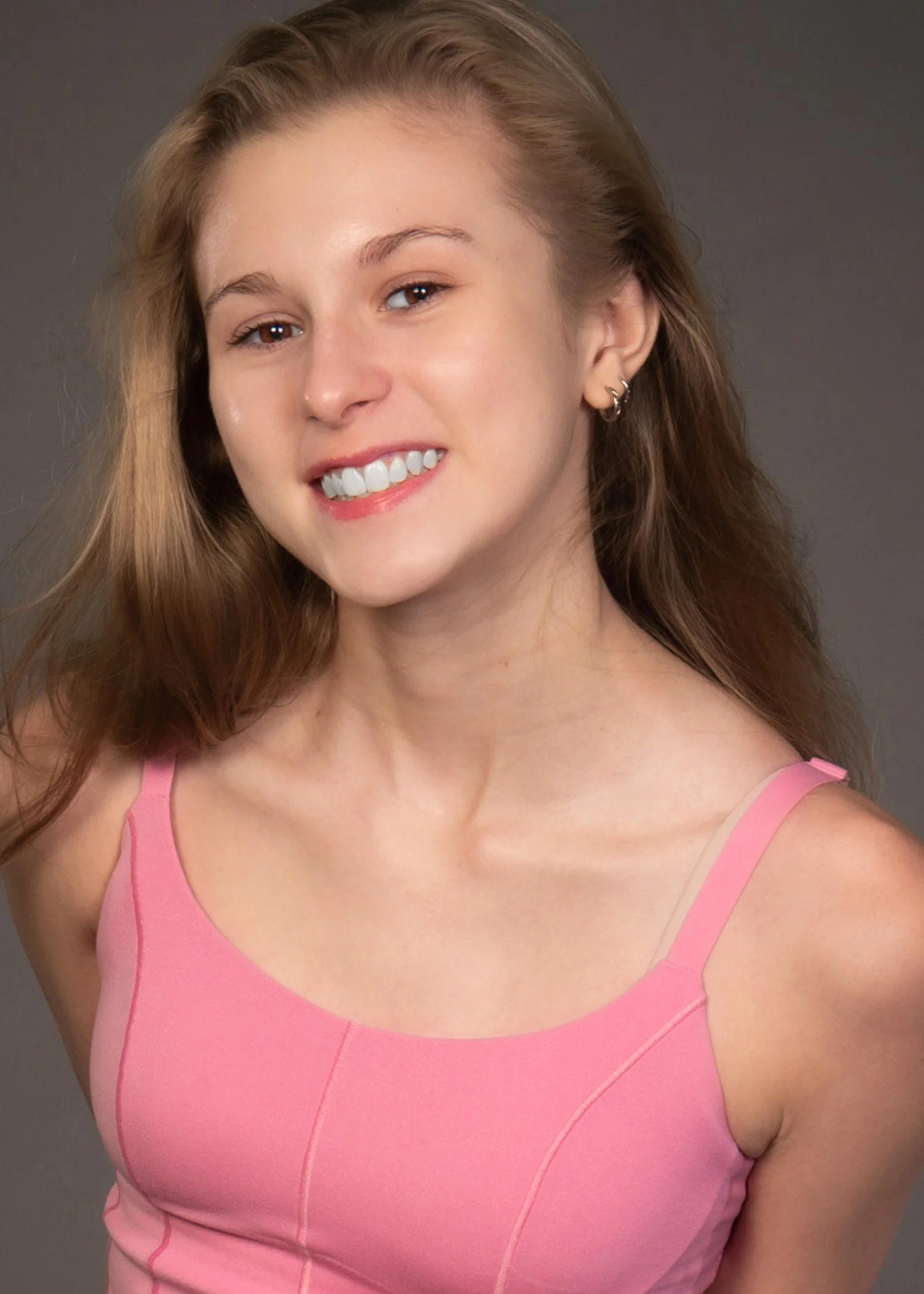 A young woman with long, wavy dirty blonde hair wearing a pink sleeveless top, smiling and showing her teeth, with earrings, against a plain gray background.