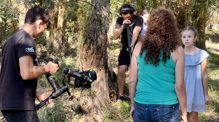 A filmmaking crew captures a scene in a wooded area with three women and a teenage boy operating camera equipment.