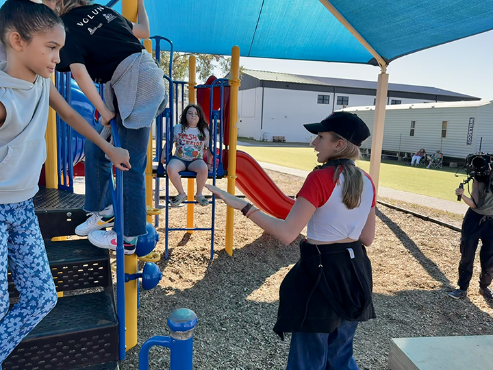 A group of children and a woman at a playground, with one girl sitting on a playground structure, other kids climbing, and a girl holding a microphone, with a person filming nearby.