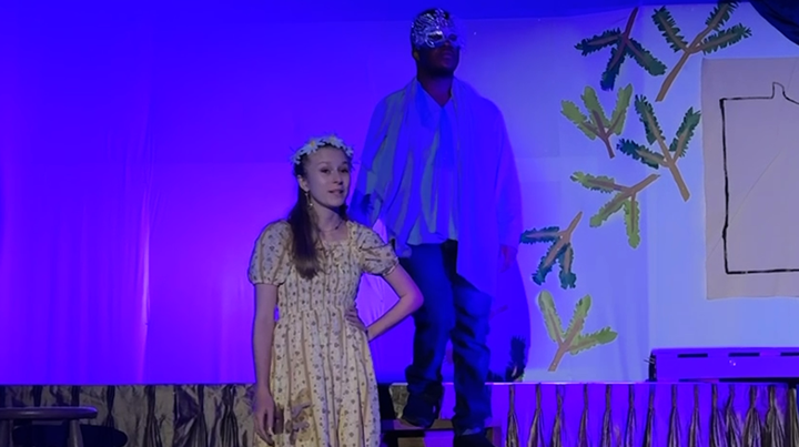 Two children are on stage during a school play or performance, with a painted backdrop depicting trees and a window. One girl is wearing a yellow dress and a flower crown, and the boy is dressed in a costume with a blue robe and a mask.
