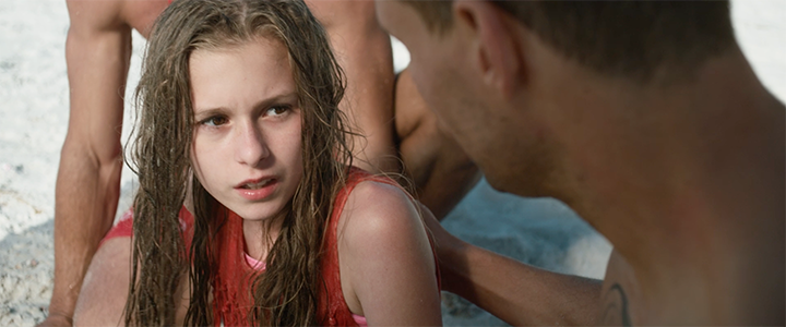 A young girl with wet hair and a distressed expression talking to a man on a beach.