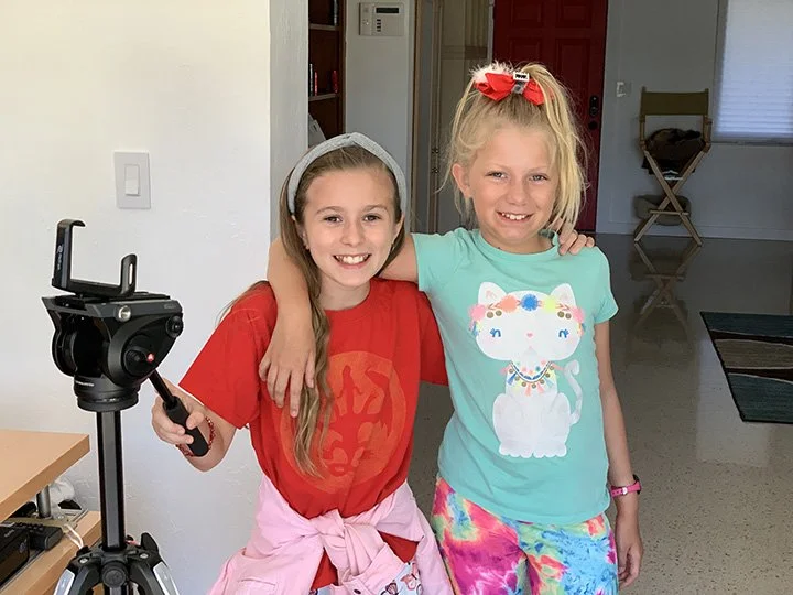 Two young girls standing indoors, smiling at the camera. One girl wears a red shirt and pink jacket tied around her waist, holding a camera on a tripod. The other girl wears a turquoise shirt with a cartoon cat and colorful shorts, with her arm around the first girl.