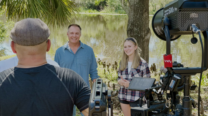 A man and a woman are being filmed outdoors by a camera operator near a pond surrounded by trees.