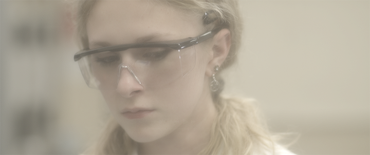 Close-up of a woman wearing safety glasses and earrings, with a focused expression.